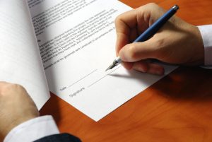 Contract with a hand holding a pen signing it where by the salesperson got their client to agree.